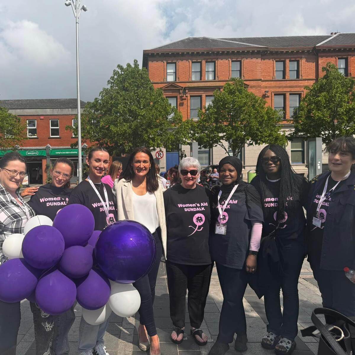 A group of women from Dundalk Women's Aid in black T-shirts with purple writing. Purple balloons in the foreground and Senator Erin McGreehan in the middle with a cream jacket and dark jeans. In the Market Square of Dundalk