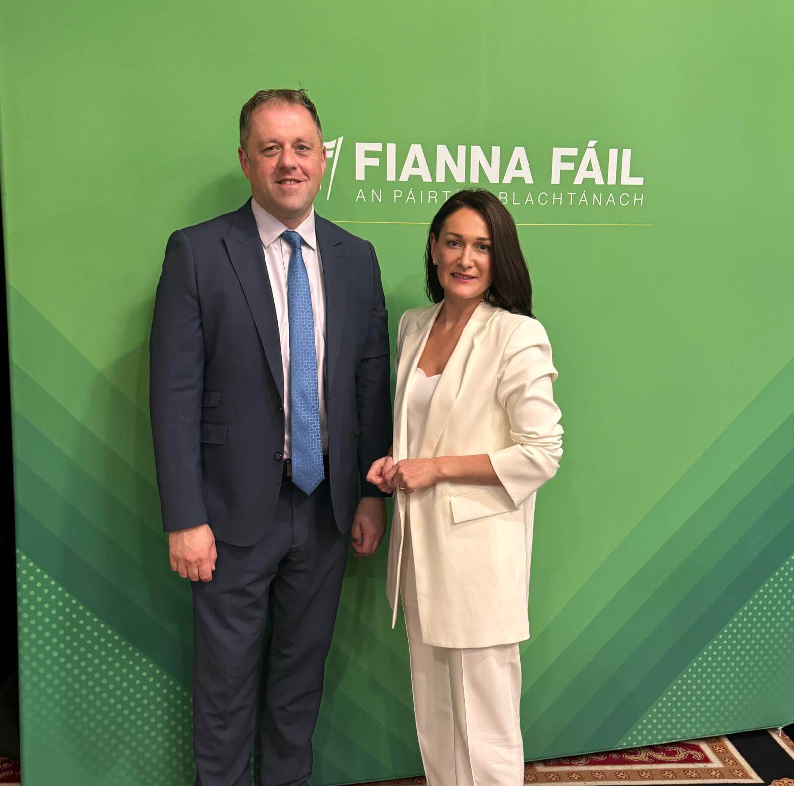 Senator Erin McGreehan standing to the right of Minister Thomas Byrne. Erin is wearing a cream suit and Thomas is wearing a dark suit, white shirt and blue tie. They are standing infront of a green Fianna Fail poster.