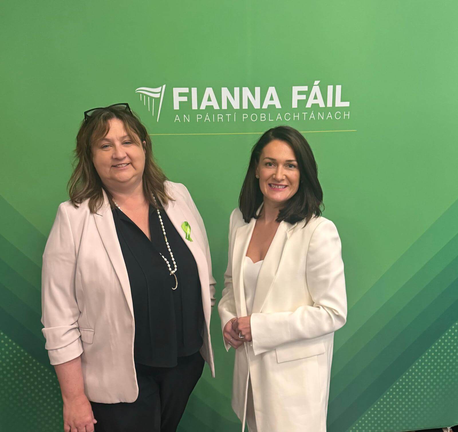 Photo of Minister for Disability Anne Rabbitte and Senator Erin McGreehan standing infront of a green Fianna Fáil poster. Erin is wearing a cream suit and Anne is wearing a cream blazer and black top.